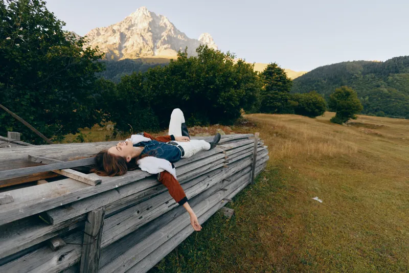 Woman lying on a wooden fence in a meadow with a mountain view, relaxing in nature among trees and open countryside, wearing casual jacket for a peaceful outdoor escape.