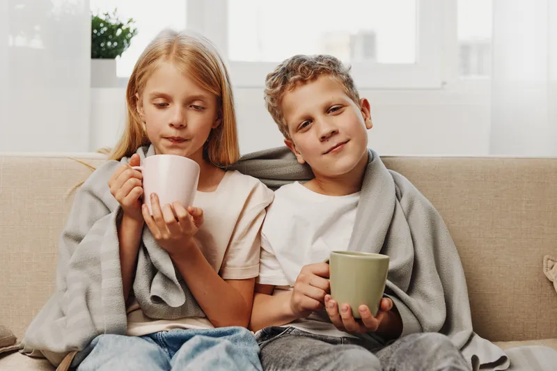 Two children wrapped in cozy blankets sit on a sofa holding ceramic mugs, enjoying a calm moment in a bright living room, symbolizing comfort, warmth, and sibling bonding.