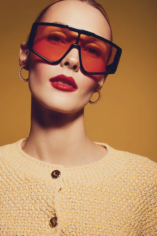 Stylish woman with oversized orange sunglasses and red lipstick poses against yellow background. She wears gold hoop earrings and a textured beige sweater. Fashion and beauty concept.