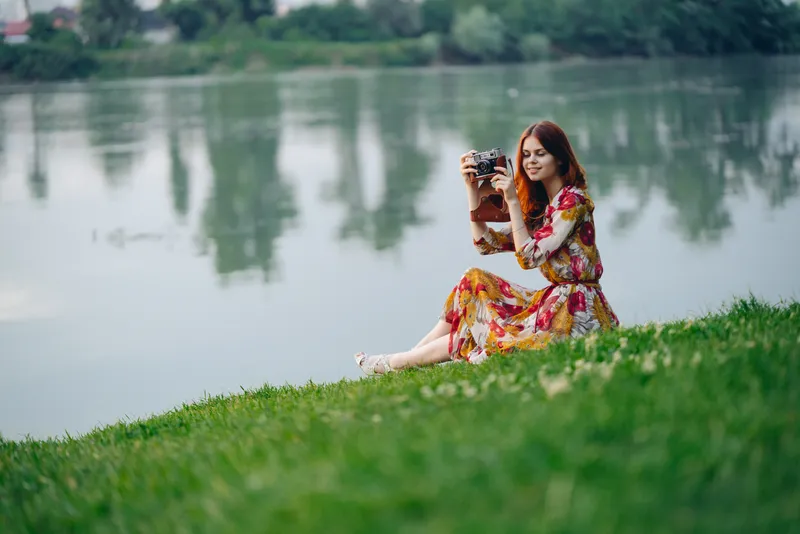 Женщина на открытом воздухе в платье с фотоаппаратом в руках на берегу озера