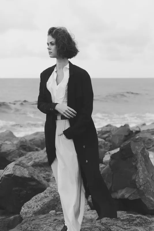 Fashion model stands on a rocky coastline by the sea in a long dark blazer over white trousers, monochrome tones emphasize elegance and minimalist style, wind swept pose conveying timeless confidence