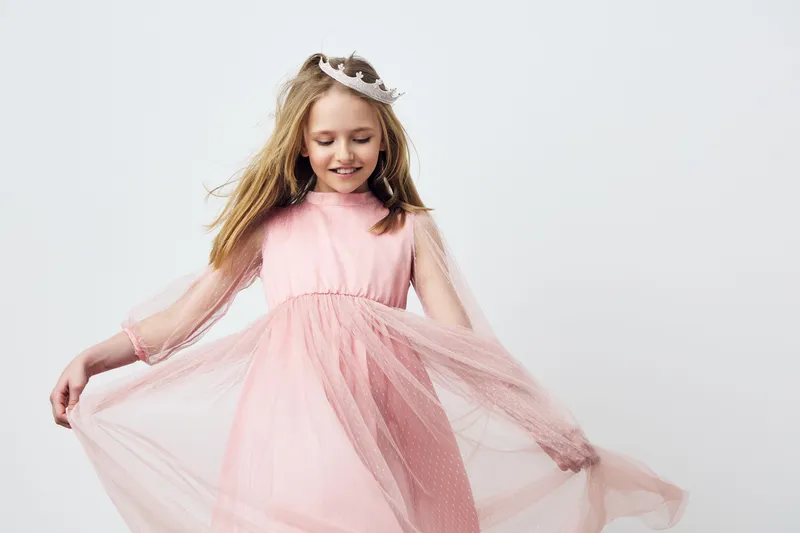 A joyful young girl in a pink dress with tulle, wearing a tiara, dancing gracefully against a soft, light background, expressing happiness and innocence