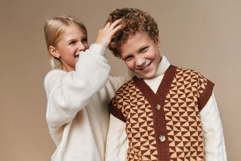 Two smiling children share playful affection as a girl gently tousles a boy's curly hair, both dressed in cozy knit sweaters against a warm studio backdrop.