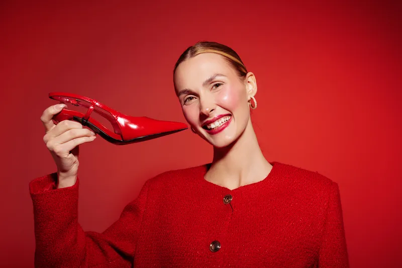 Smiling woman in red jacket holds shiny red high heel shoe near face on red background. Concept of fashion, style, glamour, and confident female model with bright makeup and accessories.