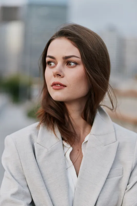Young woman in light gray blazer showing thoughtful emotion on urban background with muted cityscape vibes, natural makeup, and brown hair
