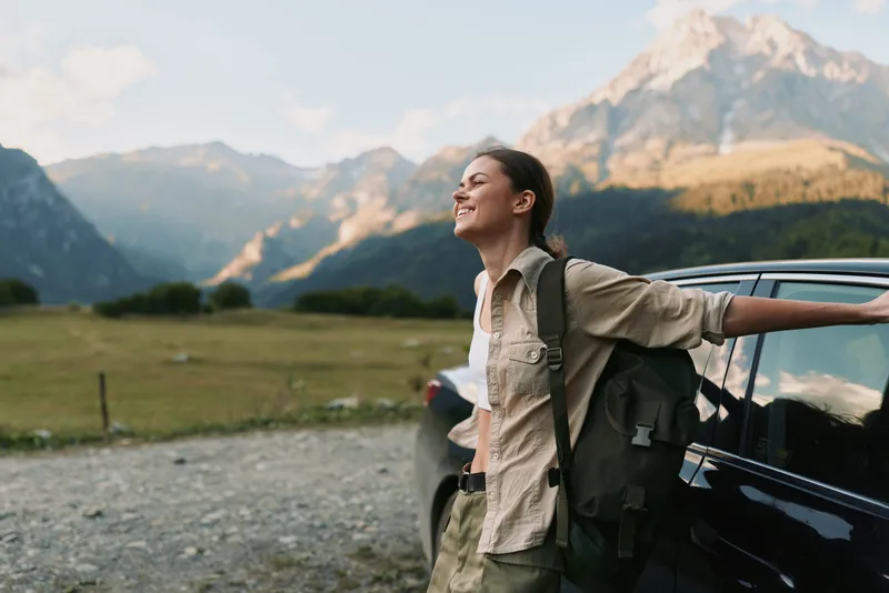 Young woman with a backpack leans against a car in a mountain valley, savoring fresh air and a feeling of freedom under clear blue skies