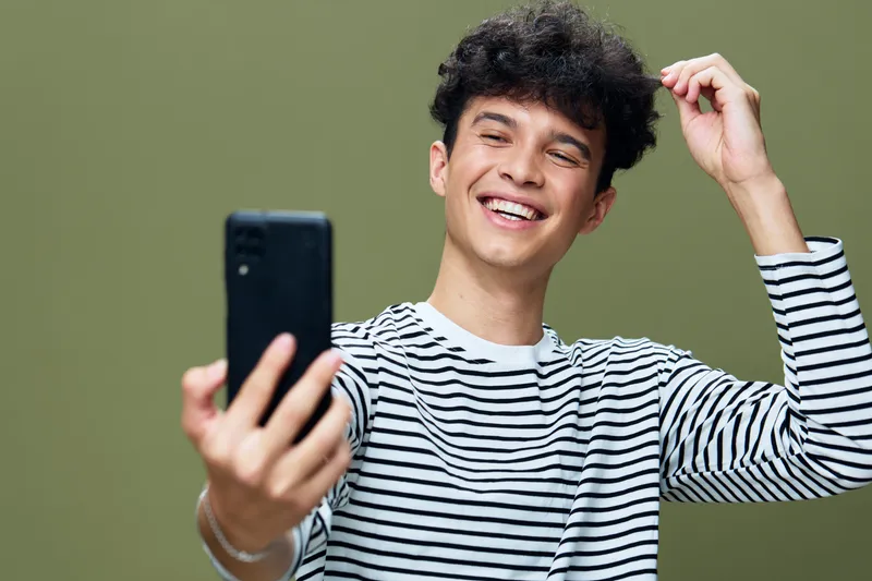 smiling young man taking selfie with smartphone, curly hair, striped shirt, bright green background, casual style, joyful expression, isolated portrait