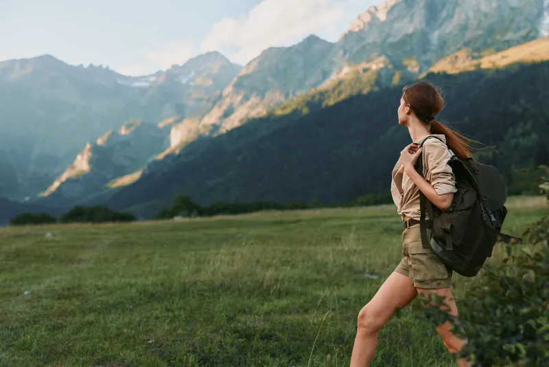 A hiking woman with a backpack strides across a green meadow, mountains rise in the distance, conveying outdoor adventure and energetic optimism under a clear sky.