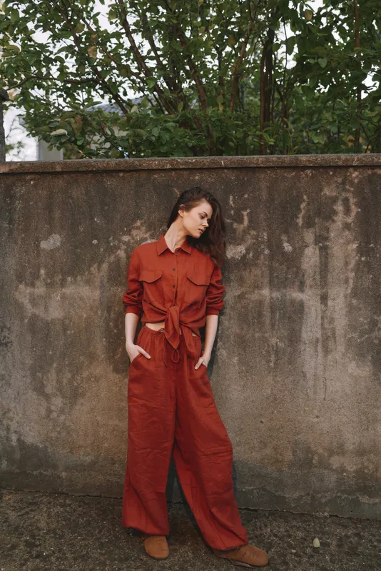 Woman in a red jumpsuit stands against a weathered wall, relaxed pose, soft natural light. Focus on fashion, streetwear, and confident everyday glamour for editorial use