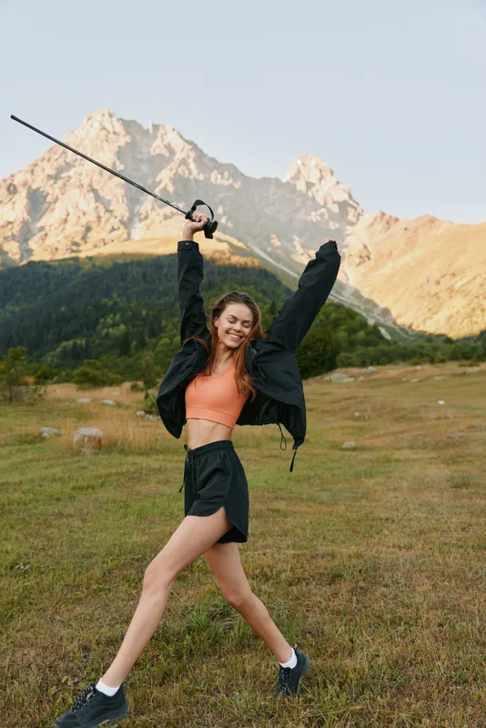 A cheerful young woman enjoys an energetic outdoor moment, raising a long stick in a wide grassy field with rugged mountains in the background, radiating freedom and vitality.