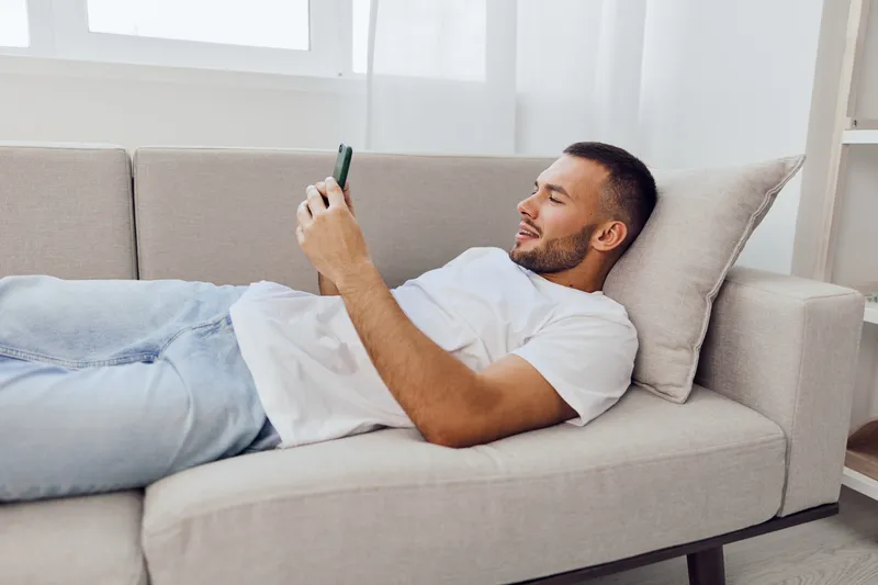 Relaxed man using his smartphone while lounging on a cozy sofa, enjoying modern technology in a minimalist living space filled with natural light.