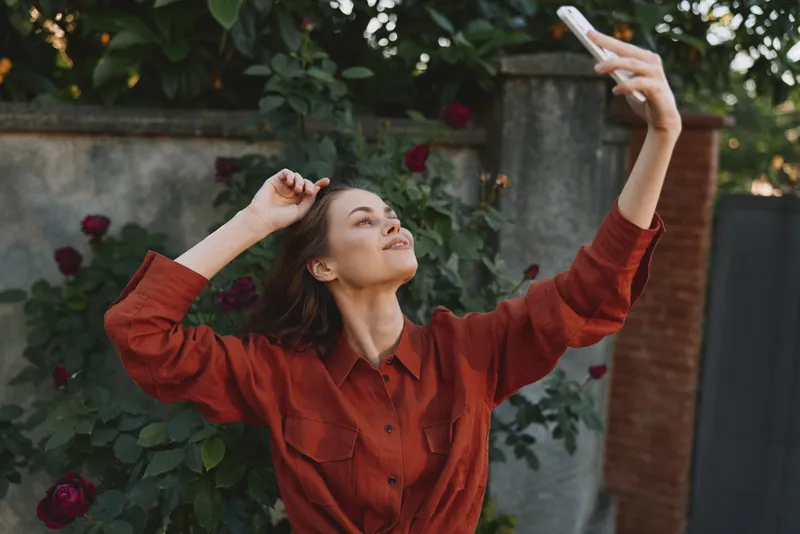 Woman lifestyle shot of a cheerful woman in a red shirt taking a selfie in a sunny garden, casual pose, candid moment, outdoor portrait