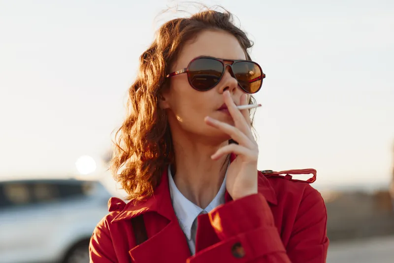 Young woman with curly hair wearing a red coat and sunglasses smokes a cigarette on an outdoor street, capturing a candid urban moment in natural light.
