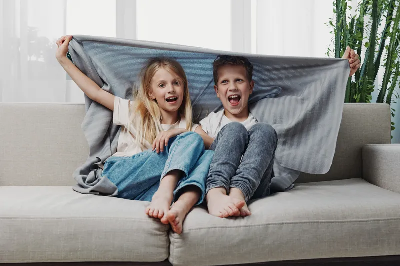 Happy children sitting on sofa covered with blanket, laughing and enjoying cozy moment in bright living room with natural light and casual clothes.