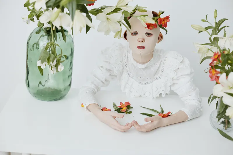 Ethereal girl in white attire surrounded by lilies on a minimalist table, expressing tranquility and softness, ideal for concepts of beauty and purity