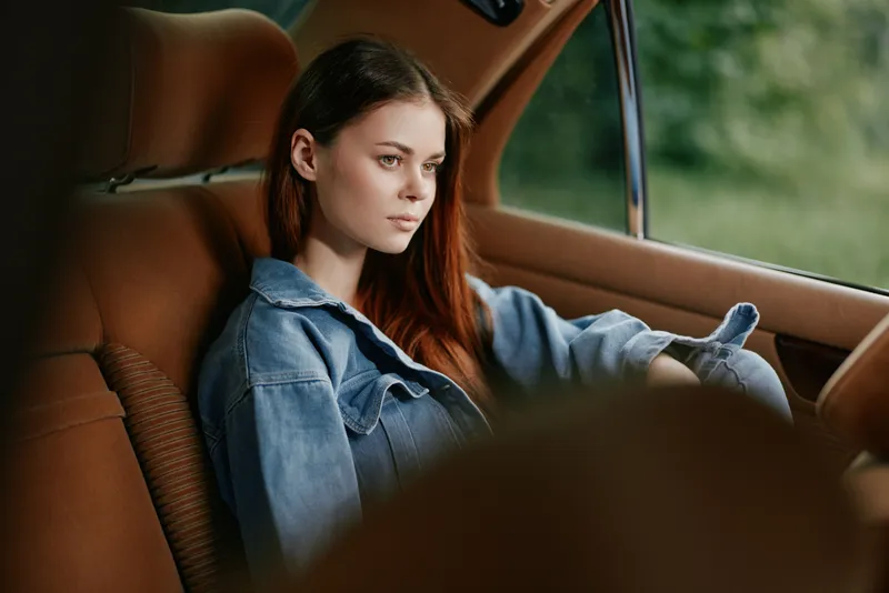 A young, stylish woman with long brown hair wearing a denim jacket, seated in a vintage car, exuding a thoughtful and dreamy expression amidst a natural backdrop.