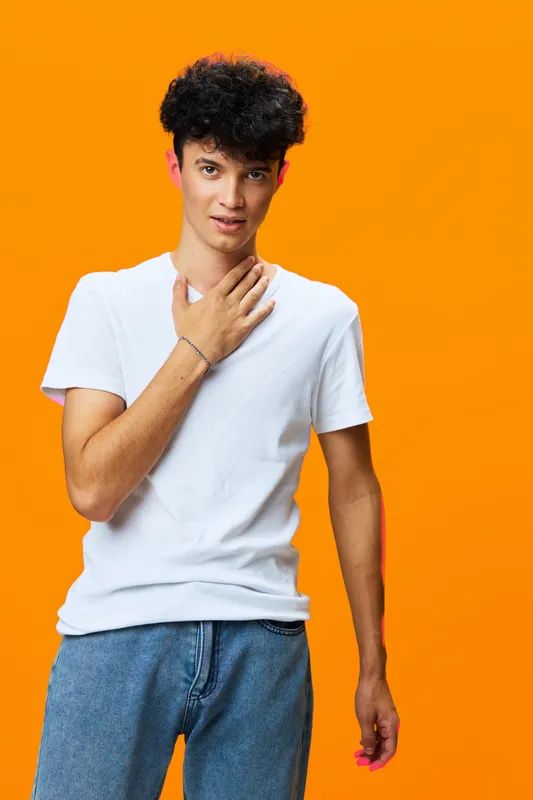 young man standing against bright orange background wearing white t-shirt and blue jeans, casual style with curly hair and relaxed posture expressing thoughtfulness