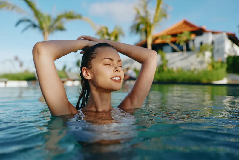 Relaxed woman enjoys swimming and sun in a tropical pool setting, eyes closed, hair wet, peaceful mood, vacation vibe, bright outdoor sanctuary, warm breeze and calm water enhance serenity