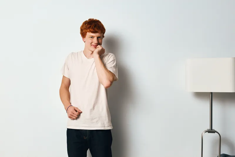Young man with red hair wearing light t shirt and dark jeans stands by white wall with floor lamp indoors. Casual style, smiling, relaxed pose, modern minimalistic interior background.