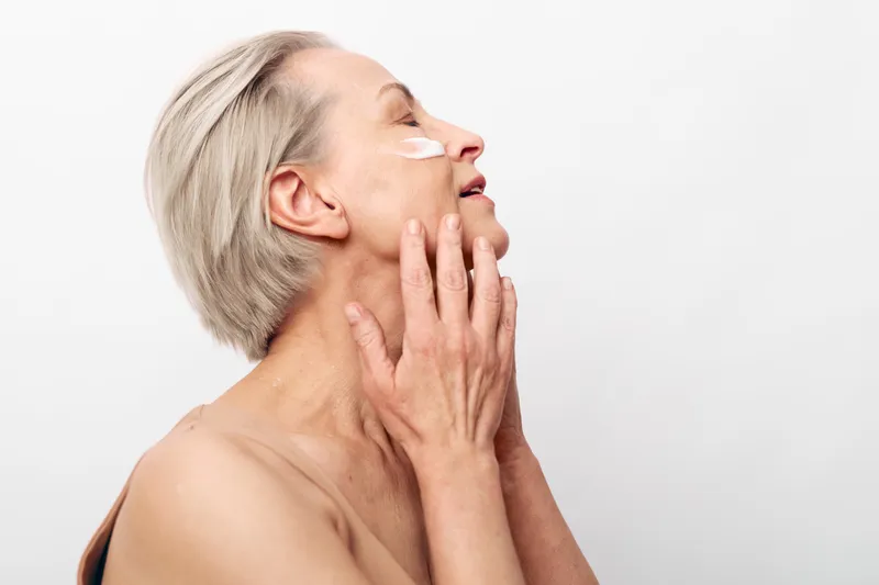 Mature woman with short grey hair applies skincare cream under eyes, gently touching face with hand. Concept of aging skin care, beauty routine, self-care, and healthy glow on white background.