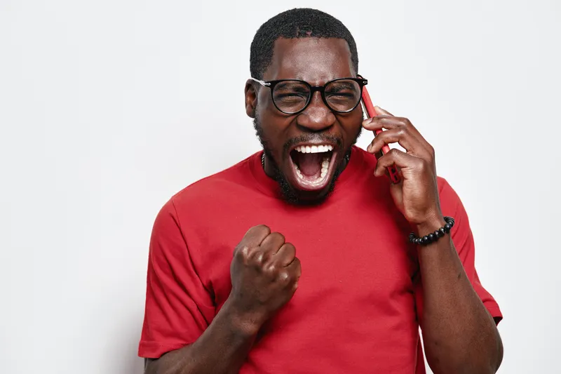 African American man with dark skin wearing glasses and red shirt talking on phone and celebrating success with fist pump and open mouth expression.