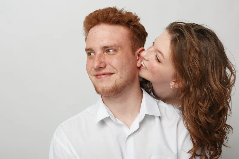 Young couple in white shirts share a tender moment, woman kissing man on cheek. Studio portrait with neutral background, concept of love, affection, and relationship happiness.