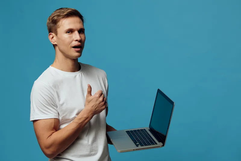 Confused young man holding open laptop with blank screen, gesturing with his hand, expressing surprise or doubt, isolated on blue background, casual white t-shirt, studio portrait. People emotion conc