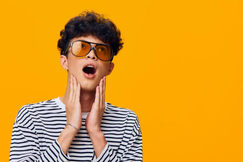 surprised young man wearing striped shirt and yellow sunglasses expressing amazement on bright orange background with curly hair in casual style