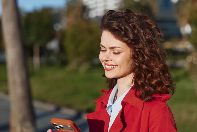 Smiling woman with curly hair wearing red coat using smartphone outdoors on sunny day. Concept of communication, technology, and happiness in urban environment.