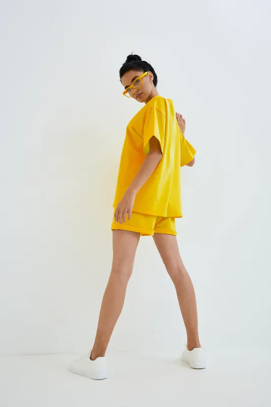 Young woman wearing bright yellow casual outfit and glasses stands confidently against plain white background, showcasing modern summer style and vibrant colors.