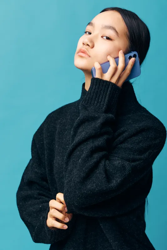 Young woman wearing black sweater holds smartphone to ear with serious expression on blue background. Casual modern style and communication concept with technology and mobile device.