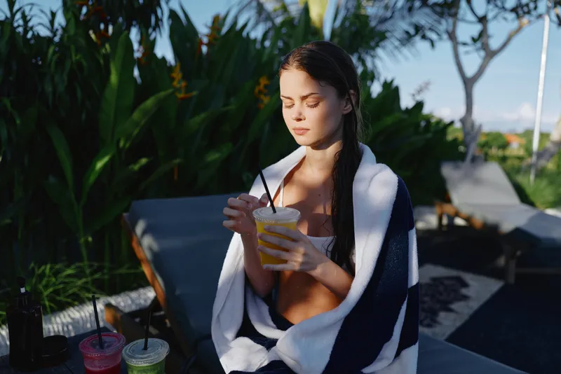 Cheerful woman outdoors, poolside moment, sipping a smoothie from a cup, wrapped in a towel, sunlit scene with tropical plants, relaxed mood