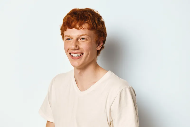 Young man with red hair smiling and looking away in white t shirt against light background for portrait and happy expression.