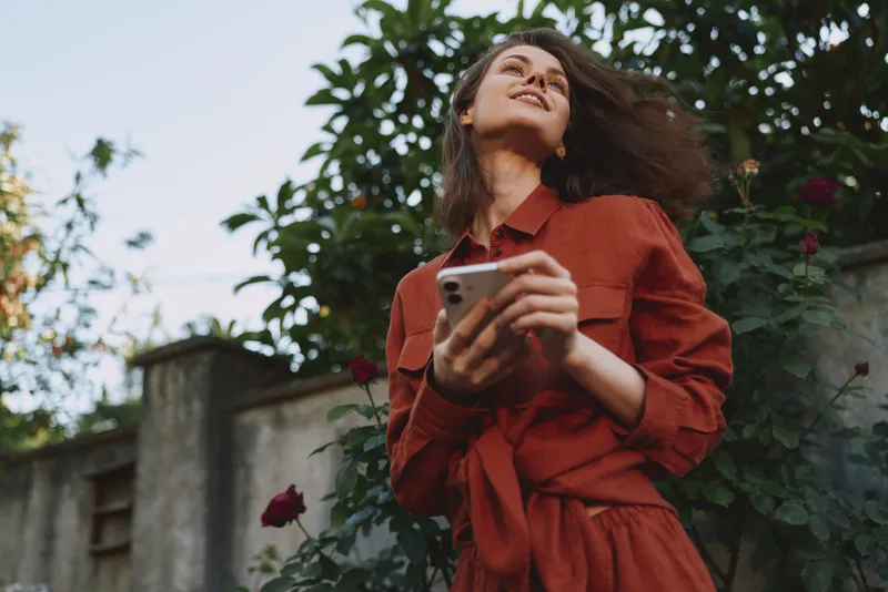A woman in a red dress stands outdoors, head tilted back, phone in hand, bathed in warm sunlight. A candid lifestyle moment capturing relaxed confidence and everyday joy