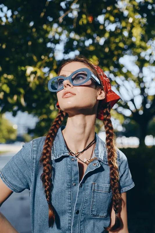 Modern boho-western denim woman wearing oversized sunglasses and a red bandana, styled in casual streetwear with braided hair under film color sunlight.