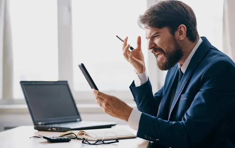 Эмоциональный мужчина на работе испытывает стресс и раздражительность, кричит, сидя в костюме перед ноутбуком