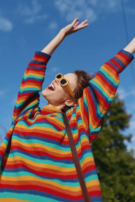 woman rainbow sweater sunglasses joyful outdoor young adult arms raised sunny. Smiling twenty something female in a colorful striped knit and retro yellow frames, casual outfit celebrating in park