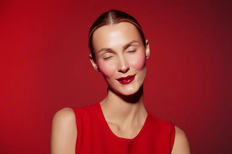 Young woman with closed eyes and red lipstick smiles softly against red background. She wears a sleeveless red top. Concept of beauty, makeup, fashion, and confidence.