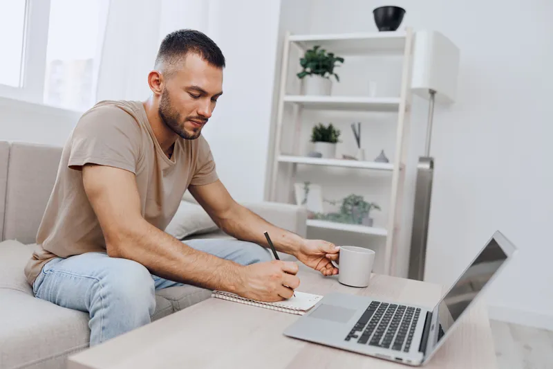 Focused man writing notes at home while enjoying a warm beverage beside his laptop, showcasing a modern and cozy workspace aesthetic perfect for productivity.