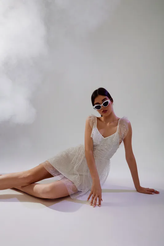 Young woman in a white dress and sunglasses poses on the floor in a studio with soft smoke. Concept of woman beauty fashion and modern style in photography.