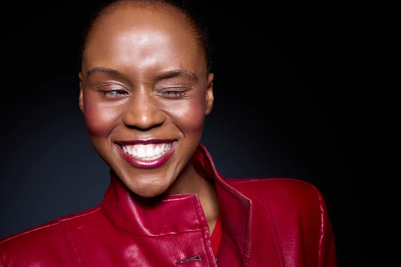 Close-up of a smiling black woman with dark skin wearing a red leather jacket winking. Studio portrait with dark background. Concept of confidence, happiness, and fashion style.