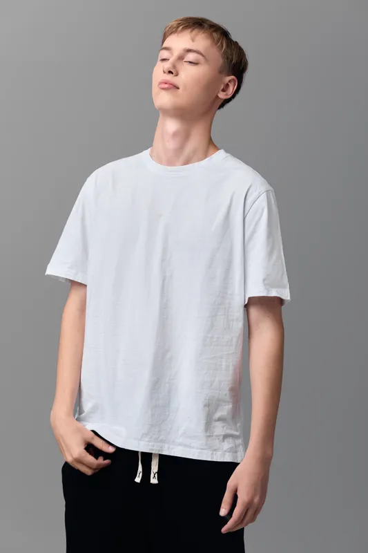 Young man in a white t shirt stands in a neutral studio, adopting a relaxed pose that conveys casual style and everyday wear, against a minimalist backdrop for a clean, versatile fashion shot.