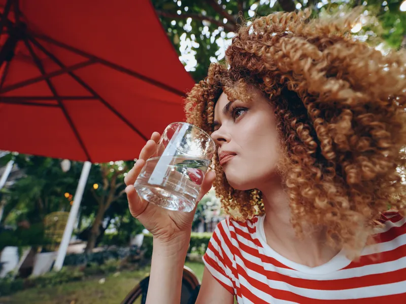 Portrait of a young woman with curly hair drinking water from a glass outdoors beneath a red umbrella, wearing a striped shirt, capturing a calm refreshment moment.