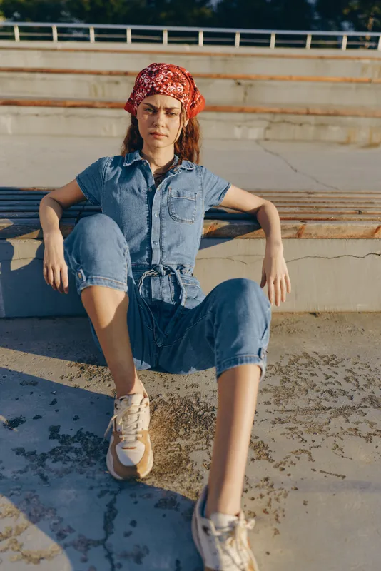 Lifestyle woman in modern boho-western denim street style wearing bandana and sneakers, relaxed pose outdoors, warm film color tones capturing casual mood.