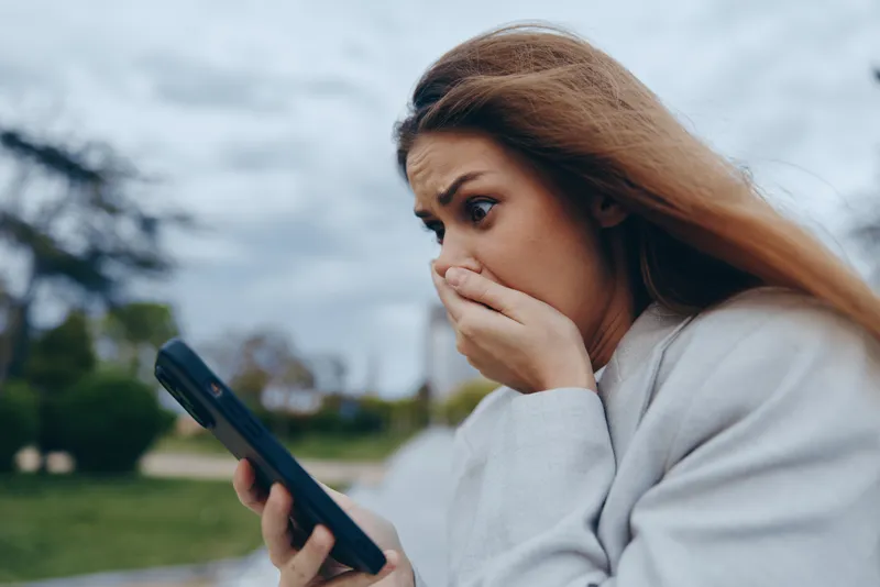 A surprised businesswoman covering her mouth while looking at her phone, showcasing modern work-life dynamics and emotional reaction outdoors.