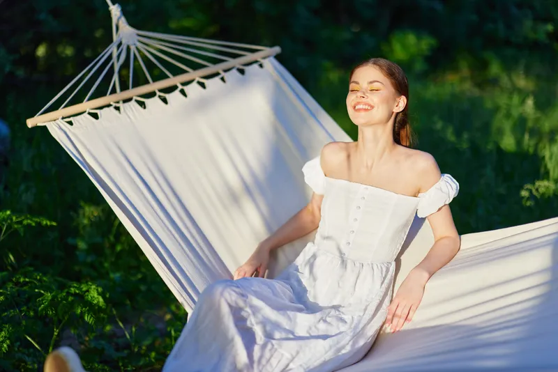 Young woman enjoying sunlight in a hammock, wearing a white summer dress, smiling happily in a green outdoor setting during daytime