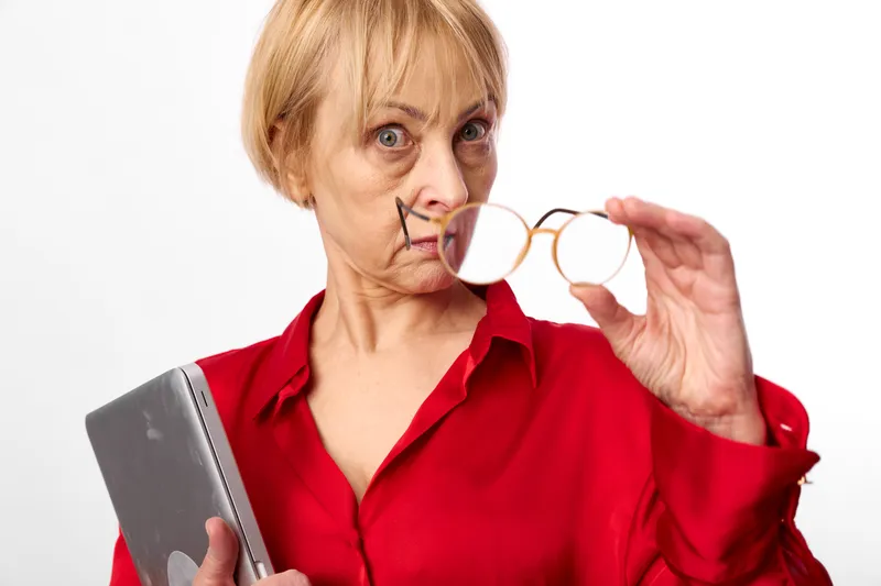 Portrait of elderly woman in studio, holding eyeglasses near face, checking vision and reading glasses, confident thoughtful expression, red blouse on plain gray background