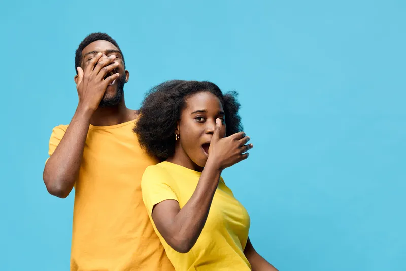 Young Black couple expressing surprise with playful gestures against a bright blue background, showcasing joy and spontaneity in vibrant yellow outfits