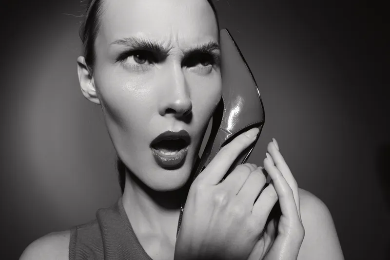 White woman with light skin holds high heel shoe to ear like phone with surprised expression. Black and white portrait with dramatic lighting and fashion concept.