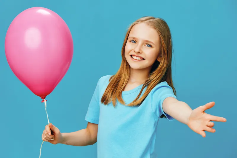Cheerful girl holding a pink balloon with an outstretched hand against a vibrant blue background, embodying joy, youth, and playfulness in a colorful setting
